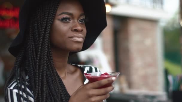 Close-up van sexy Afrikaanse Amerikaanse vrouw in zwarte hoed en gestreepte vogue jurk met koele cocktail in bar. Hete zomer dag refreshness — Stockvideo