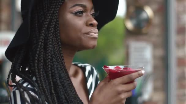 Close-up de sexy e atenciosa senhora afro-americana em chapéu preto e vestido de voga listrado com coquetel legal no bar. Refrescamento quente do dia de verão — Vídeo de Stock