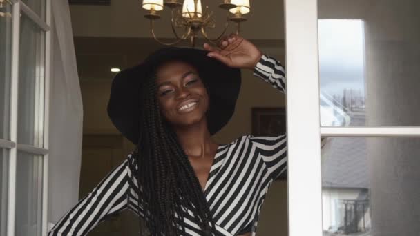Close-up van elegante sexy Afro-Amerikaanse vrouwelijke model in zwarte hoed en gestreepte jurk glimlachen op de luxeappartementen. Vogue fashion concept — Stockvideo