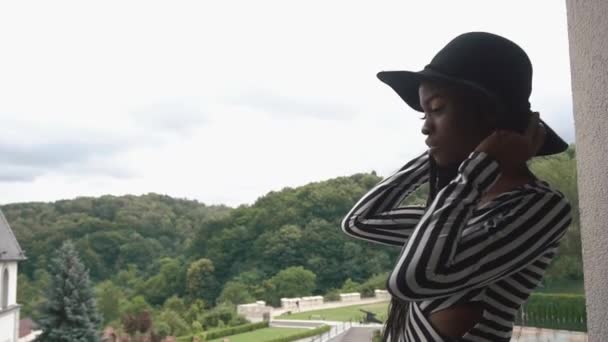 Elegante senhora afro-americana de chapéu preto em pé na varanda de seus apartamentos e observando a vista — Vídeo de Stock