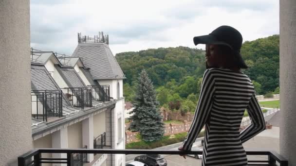 Элегантная африканская американка в черной шляпе и модное полосатое платье, стоящее на балконе ее роскошных апартаментов и смотрящее на вид — стоковое видео