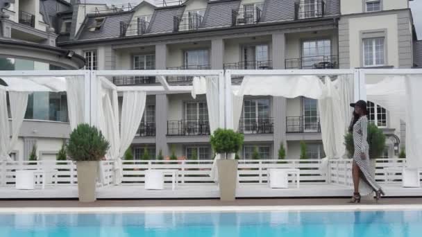 Vue latérale de sexy élégant modèle féminin afro-américain en robe de cocktail et chapeau noir se promène à la piscine de luxe à l'hôtel au ralenti — Video