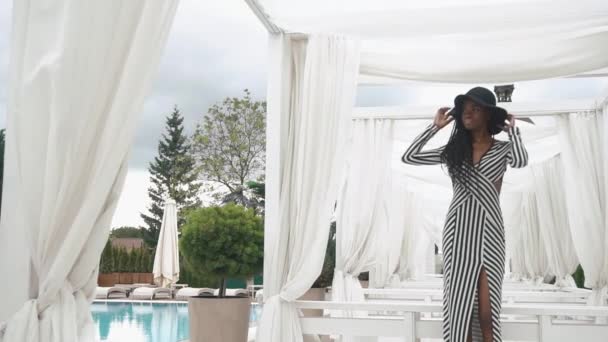 Afro-amerikai hölgy elegáns csíkos koktél ruha és fekete kalapban pózol a medencénél, luxus hotel háttérben. — Stock videók