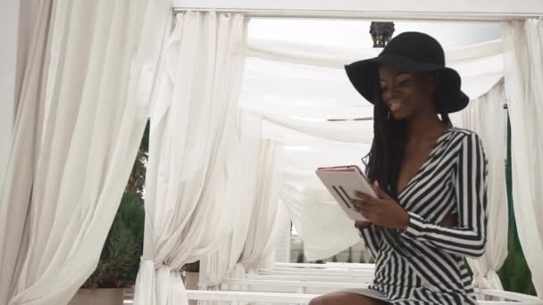 Okouzlující afroamerické dáma v krátké koktejlové šaty a černý klobouk pomocí svého tabletu a usmívá se. Luxusní hotel venkovní pozadí — Stock video
