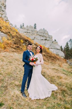 Şık mavi takım elbiseyakışıklı damat backround olarak kayalar ile pastoral açık arazi üzerinde gül buket tutan beyaz giyinmiş gelin kucaklayan