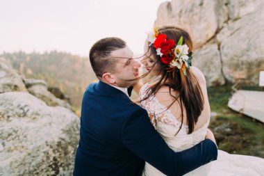 Şık mavi takım elbiseli sevilen damat arka yuvarlak olarak görkemli dağ kayalık manzara üzerinde sevimli baş çelenk ile romantik beyaz giyinmiş gelin tutarak. Geri görünüm