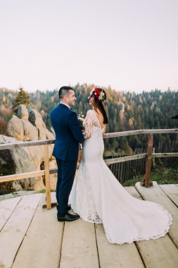 Şık mavi takım elbiseli zarif damat sevgiyle arka plan olarak görkemli dağ kayalık manzara ile ahşap platform üzerinde duran onun büyüleyici beyaz giyimli gelin tutarak. Geri görünüm