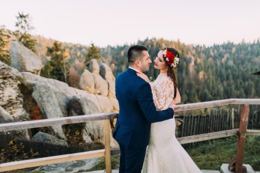 Şık mavi takım elbiseli zarif damat sevgiyle arka plan olarak görkemli dağ kayalık manzara ile ahşap platform üzerinde duran onun büyüleyici beyaz giyimli gelin tutarak