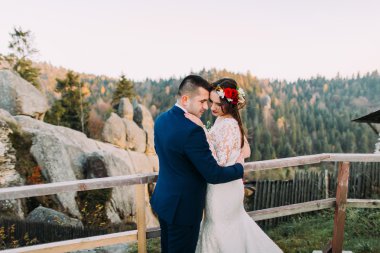 Şık mavi takım elbiseli zarif damat sevgiyle arka plan olarak inanılmaz dağ kayalık manzara ile ahşap platform üzerinde duran onun büyüleyici beyaz giyimli gelin tutarak. Geri görünüm