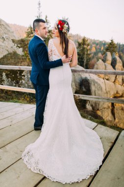 Zarif damat şık mavi takım sevgiyle arka plan olarak görkemli dağ kayalık manzara ile ahşap platform üzerinde duran muhteşem uzun beyaz elbise onun büyüleyici gelin tutarak. Geri görünüm