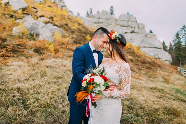 Handsome groom in stylish blue suit embracing white dressed bride holding bouquet of roses on idyllic pastoral landscape with rocks and fence as backround — Stock Photo, Image