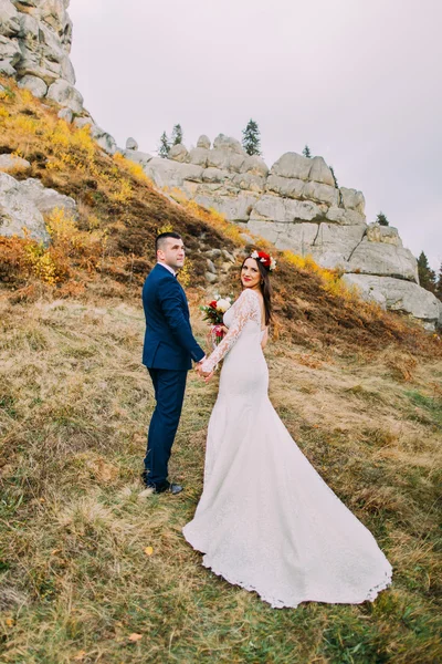 Guapo novio en elegante traje azul abrazando novia vestida blanca sosteniendo ramo de rosas en idílico paisaje pastoral con majestuosas rocas de fondo — Foto de Stock