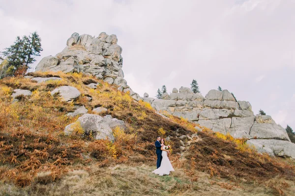 Beau marié en costume bleu élégant embrassant mariée habillée blanche tenant bouquet de roses sur paysage extérieur idyllique avec des roches merveilleuses comme backround — Photo