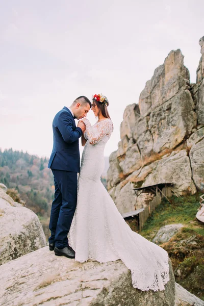 Novio guapo en elegante traje azul cariñosamente besando a su novia vestida de blanco manos en majestuoso paisaje de montaña con grandes rocas como fondo — Foto de Stock