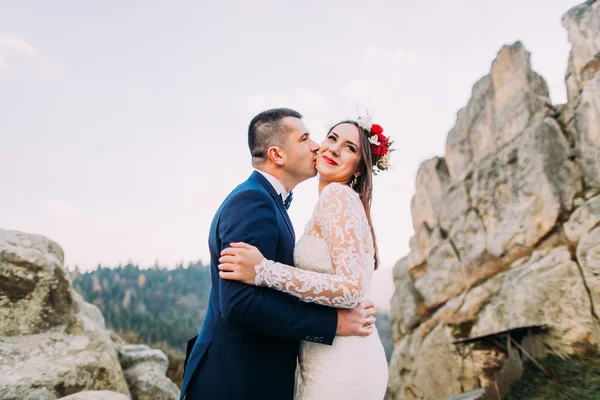 Close up view handsome groom in stylish blue suit kissing white dressed bride cute head wreath on majestic mountain rocky landscape as backround — Stock Photo, Image