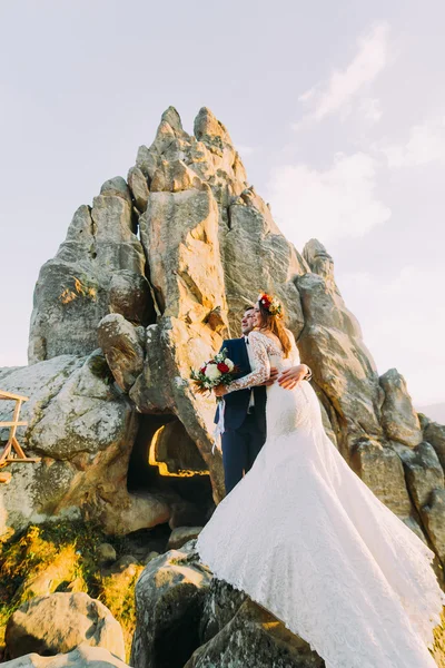 Porträtt av romantisk nygifta par i solnedgången ljus på majestätiska bergslandskap med stor stenig klippa som bakrund — Stockfoto