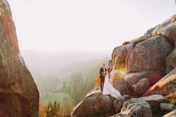 Couple de jeunes mariés romantique posant dans les lumières du coucher du soleil sur une majestueuse falaise rocheuse avec vue sur la campagne comme arrière-plan — Photo