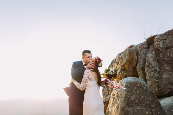 Romántico recién casado marido besar hin nueva esposa en las luces del atardecer en majestuoso acantilado de montaña rocosa —  Fotos de Stock