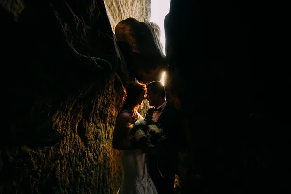 Novia hermosa romántica y elegante novio con estilo en la hendidura de roca oscura con fondo por la puesta de sol brillante — Foto de Stock