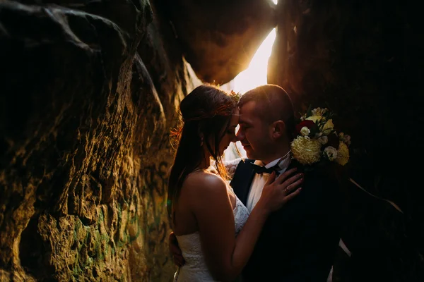 Romantik güzel gelin ve şık şık damat gün batımı parlayan arka planda karanlık kaya yarık birbirlerine tutarak — Stok fotoğraf