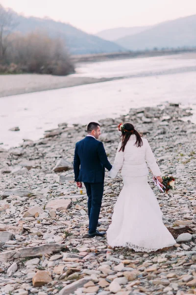 Joven pareja de novias atractiva caminando en la orilla de guijarros cerca del río de montaña. Vista trasera —  Fotos de Stock