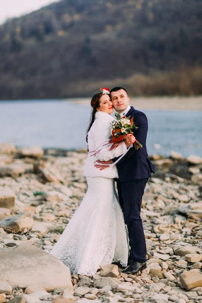 Romántica pareja de novias jóvenes abrazándose a orillas del río de guijarros con colinas forestales como fondo —  Fotos de Stock