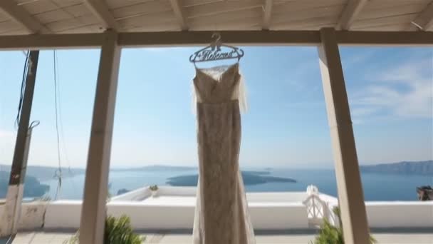 Nahaufnahme festliches Brautkleid weht im Wind auf der Terrasse mit der Aufschrift "Braut" auf dem Kleiderbügel das blaue Meer auf dem Hintergrund — Stockvideo