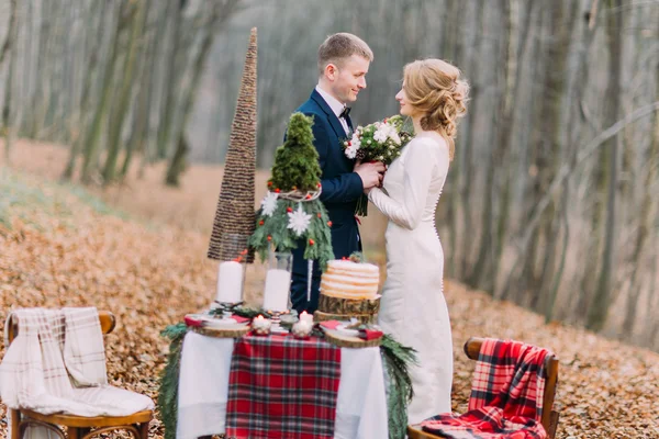 Happy young wedding couple holding hands near the decorated table for Christmas holidays in autumn forest.