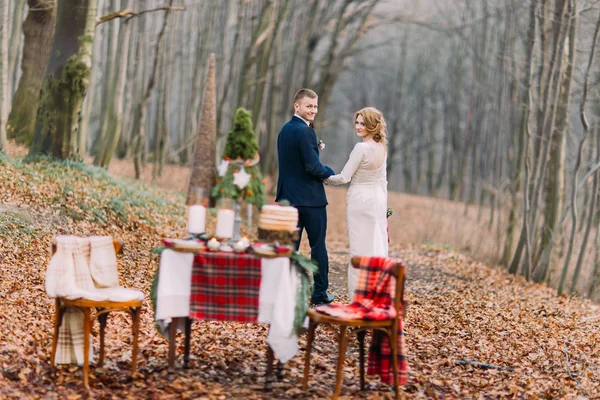 Glad ung bröllop par promenader och håller händerna nära dekorerade bordet för julhelgen i höst skog. — Stockfoto