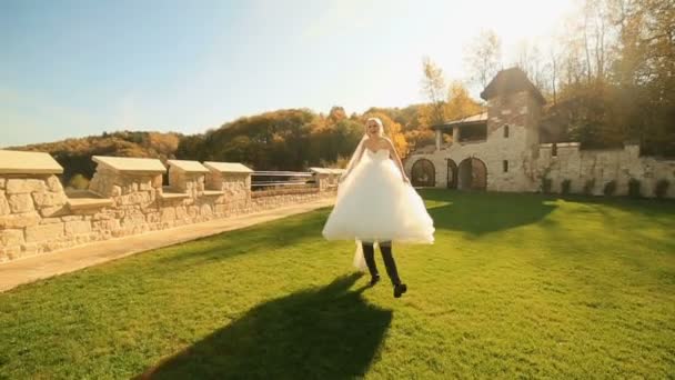 El novio feliz lleva en las manos a su adorable novia rubia. Jóvenes recién casados divirtiéndose juntos en el castillo medieval — Vídeo de stock