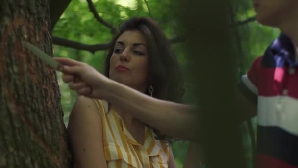 Vista laterale del giovane che intaglia un cuore con le iniziali su un albero mentre la sua donna sta guardando. Verde primavera foresta sfondo — Video Stock