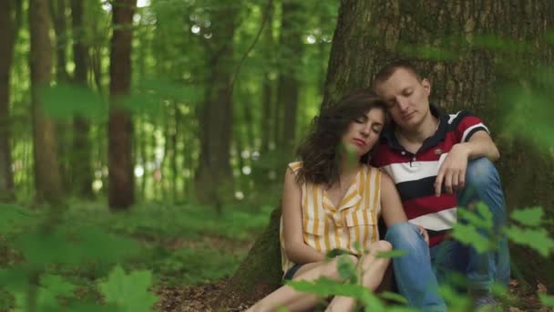 Joven feliz pareja amorosa sentado con los ojos cerrados en el árbol con el corazón tallado en él y relajante. Fondo de bosque de primavera verde — Vídeos de Stock