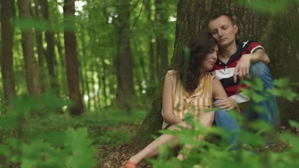 Hermosa pareja amorosa sentada con los ojos cerrados en el árbol con el corazón tallado en él y relajante. Fondo de bosque de primavera verde — Vídeos de Stock