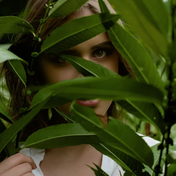 Close-up retrato de uma mulher bonita olhando através de folhas no fundo verde — Fotografia de Stock