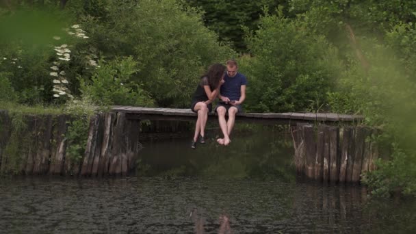 Pár ül egy fa ponton a folyó nyáron. Segítségével a digitális tábla és a feleség férj mérges a fiúról kevesebb figyelmet — Stock videók