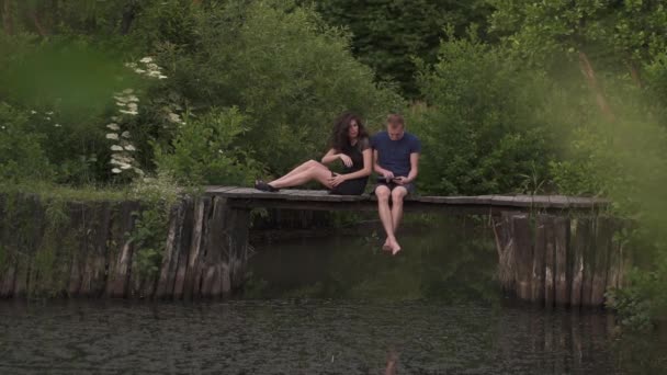 Couple assis sur un ponton en bois sur une rivière en été. Mari utilisant une tablette numérique et la femme est en colère et contrarié par moins d'attention — Video