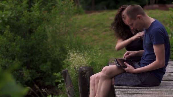 Familie paar zittend op een houten brug over een rivier in de zomer. Man met behulp van een digitaal tablet en de vrouw is boos en overstuur over minder aandacht — Stockvideo