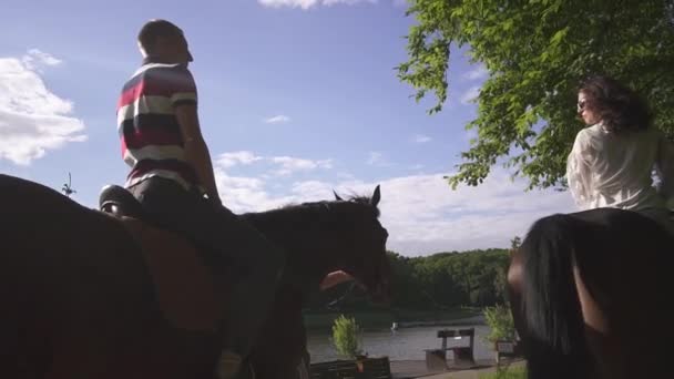 Счастливая молодая семейная пара верхом на лошадях в зеленом парке возле озера. Голубой фон неба — стоковое видео