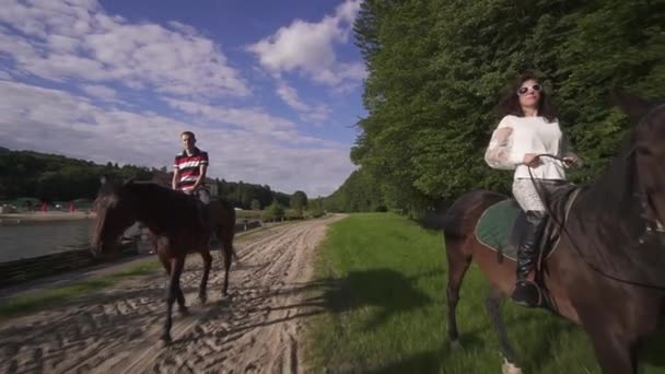 Щаслива молода сімейна пара катається на конях разом на березі річки. фон блакитного неба — стокове відео