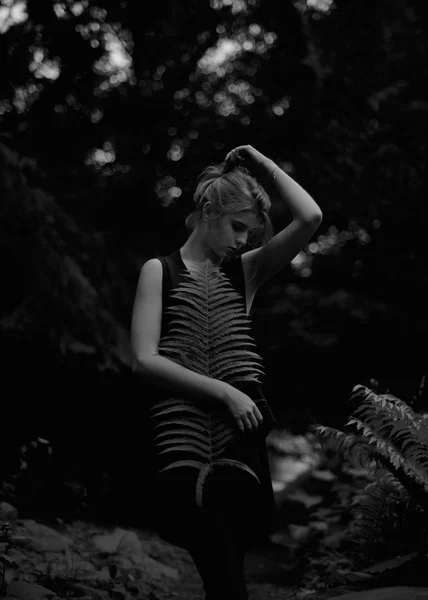 Mujer joven posando con el brazo apoyado en la cabeza entre el bosque verde, sosteniendo helecho, blanco y negro — Foto de Stock