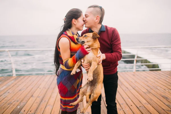 Ein junges glückliches Paar küsst sich und spielt mit dem Hund auf dem verregneten Liegeplatz im Herbst. Hintergrund Meer — Stockfoto