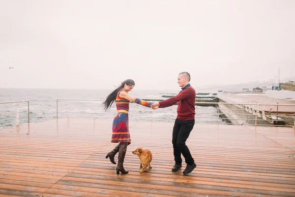 Jovem casal feliz andando com o cão e se divertindo no cais chuvoso no outono. Fundo marinho — Fotografia de Stock