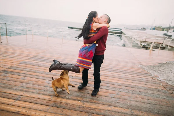 Jovem casal feliz beijando uns aos outros e brincando com o cão no cais chuvoso no outono. Fundo marinho — Fotografia de Stock