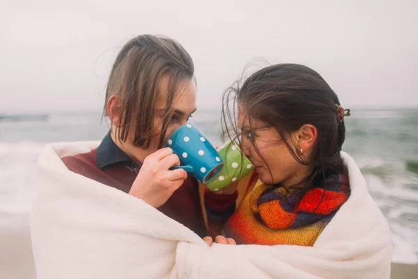 Güzel mutlu genç çift sevgiyle birbirlerine beyaz battaniye sarılmış bakmak ve sevimli renkli bardak çay içme. Arka planda kış plajı — Stok fotoğraf