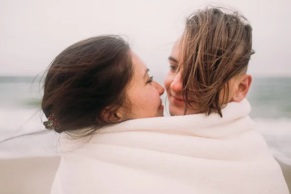 Smiling young couple softly kissing wrapped in white blanket at the winter beach. Romantic concept — Stock Photo, Image