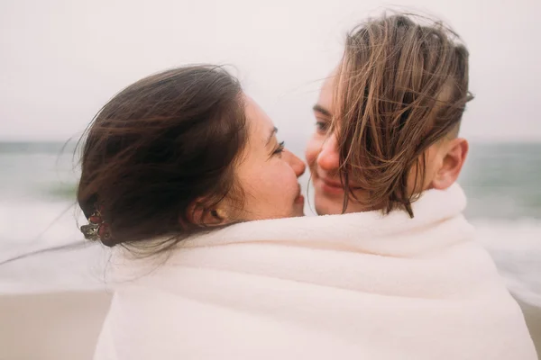 Lachend jong koppel zacht zoenen verpakt in witte deken op het winter strand. Romantisch concept — Stockfoto