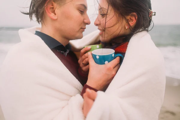 Beautiful happy young couple lovingly look at each other wrapped in white blanket with cute cups of tea. Winter beach on background — Stock Photo, Image