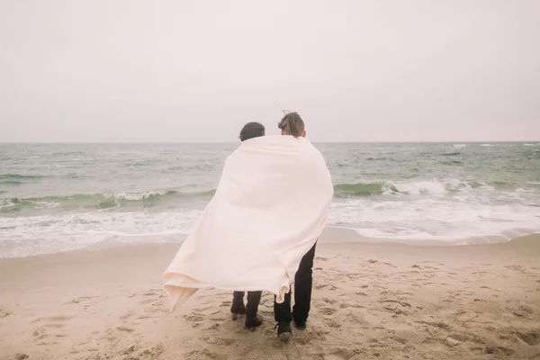 Усміхнена молода пара, загорнута в білу ковдру на зимовому пляжі. Романтична концепція — стокове фото