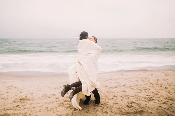 Felice giovane uomo tiene sulle mani la sua affascinante ragazza avvolta in una coperta bianca. Sfondo mare invernale — Foto Stock
