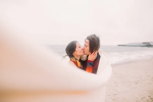 Hermosa pareja joven feliz amorosamente se miran el uno al otro envueltos en manta blanca. Mar de invierno en el fondo — Foto de Stock